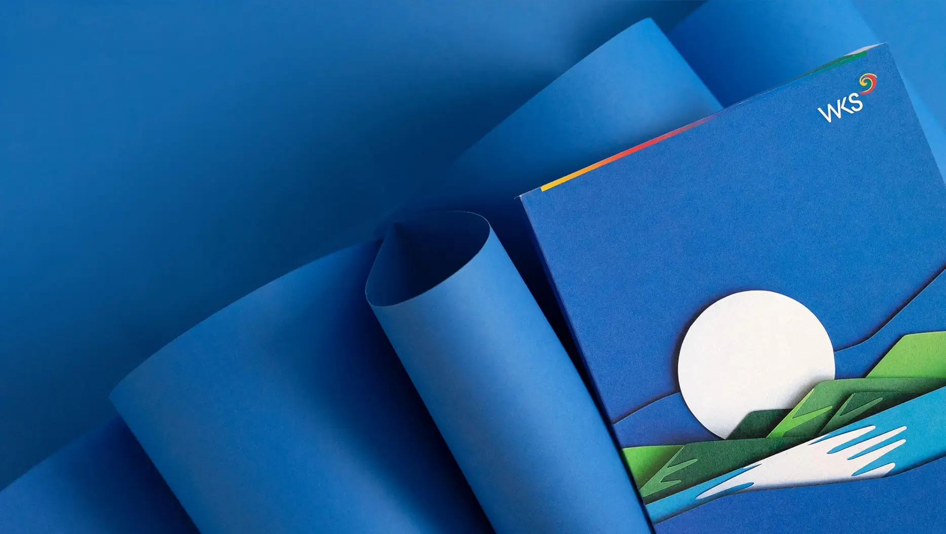 Titelseite der WKS-Umwelterklärung in Blau, mit gerollten Papierbahnen im Hintergrund — eine papierschnitt-artige Illustration aus Sonne, Bergen und Fluss ziert das Cover, oben rechts das WKS-Logo mit farbigem Akzentstreifen in Gelb, Rot und Grün.
