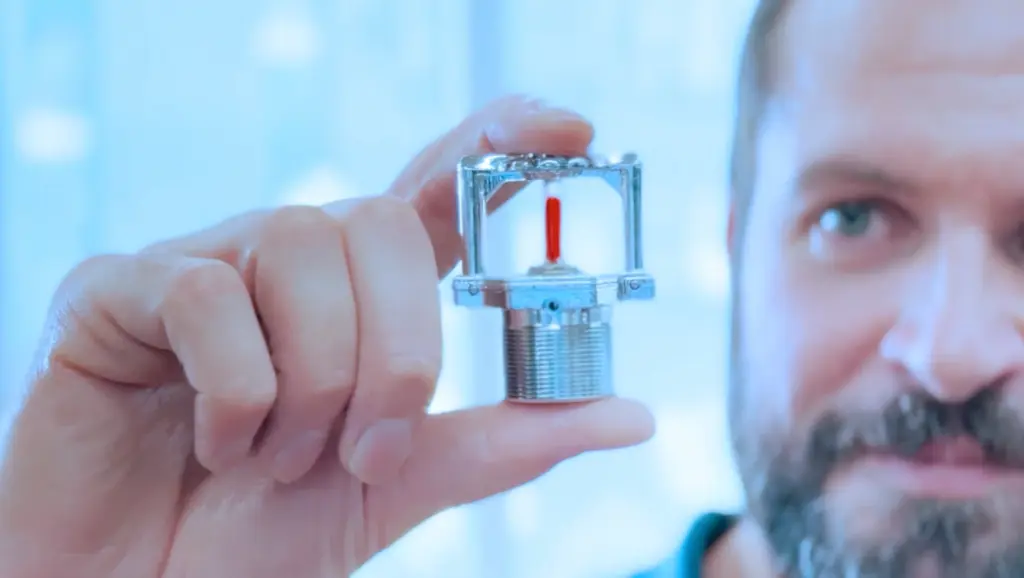 Filmstill aus dem EuroSprinkler-Unternehmensfilm — ein Mitarbeiter betrachtet konzentriert einen einzelnen Sprinklerkopf mit rotem Glaskolben in Nahaufnahme, im Hintergrund unscharf ein hell erleuchteter Produktionsraum.