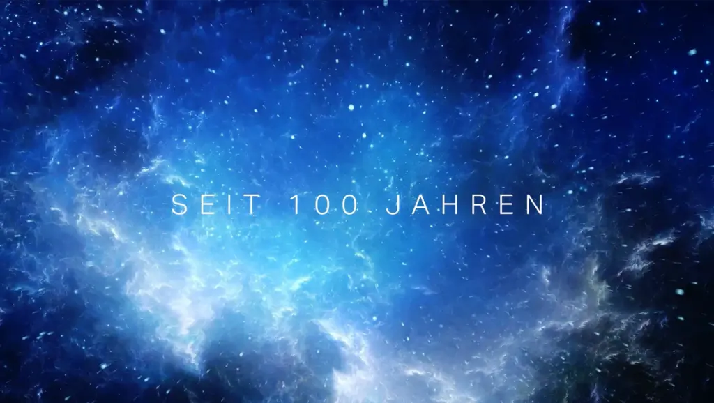 Filmstill aus dem Volkssternwarte-Köln-Imagefilm mit dem Schriftzug „Seit 100 Jahren" vor einem sternenübersäten, blau leuchtenden Nebel-Hintergrund — Szene aus dem Jubiläumsfilm zum 100-jährigen Bestehen.
