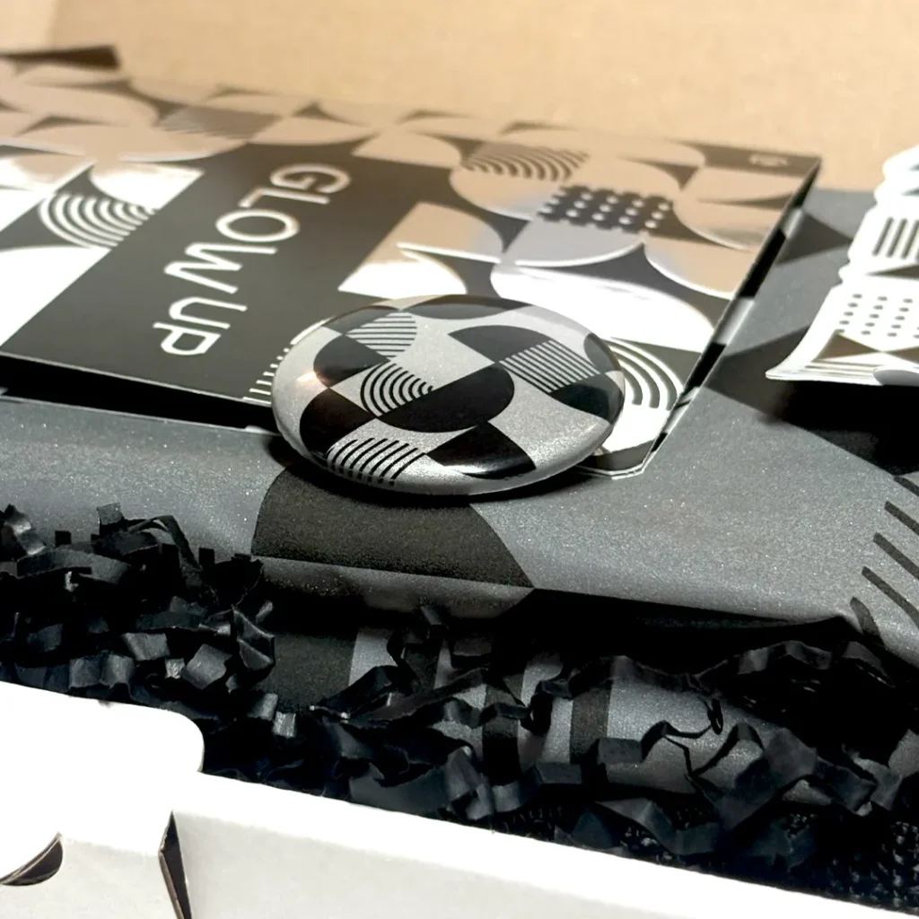 Round button with black-and-white geometric pattern from the New Year's collection “Glow Up,” lying on similarly patterned packaging. The incidence of light emphasizes the metallic reflective surface.