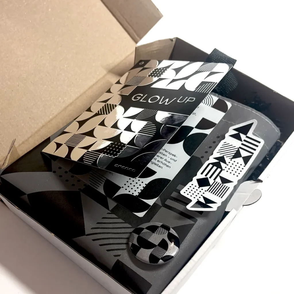 Open shipping box with the New Year's mailing “Glow Up”, consisting of a reflective card, a graphic sticker and a button with black-and-white patterns. The products are placed on decorative black filling.